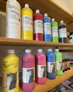 Tempera und Coache Farben in Gelb, Weiß, Magenta, Blau, Grün, Schwarz, Gold, Silber und Bronze in einem Holzregal im Kunsttherapie Atelier