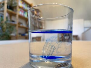 Ein Glas Wasser in einem Kunsttherapie Atelier, in das ein Tropfen blaue Tusche getropft worden ist und der sich langsam auf den Grund des Glases hinabbeuget