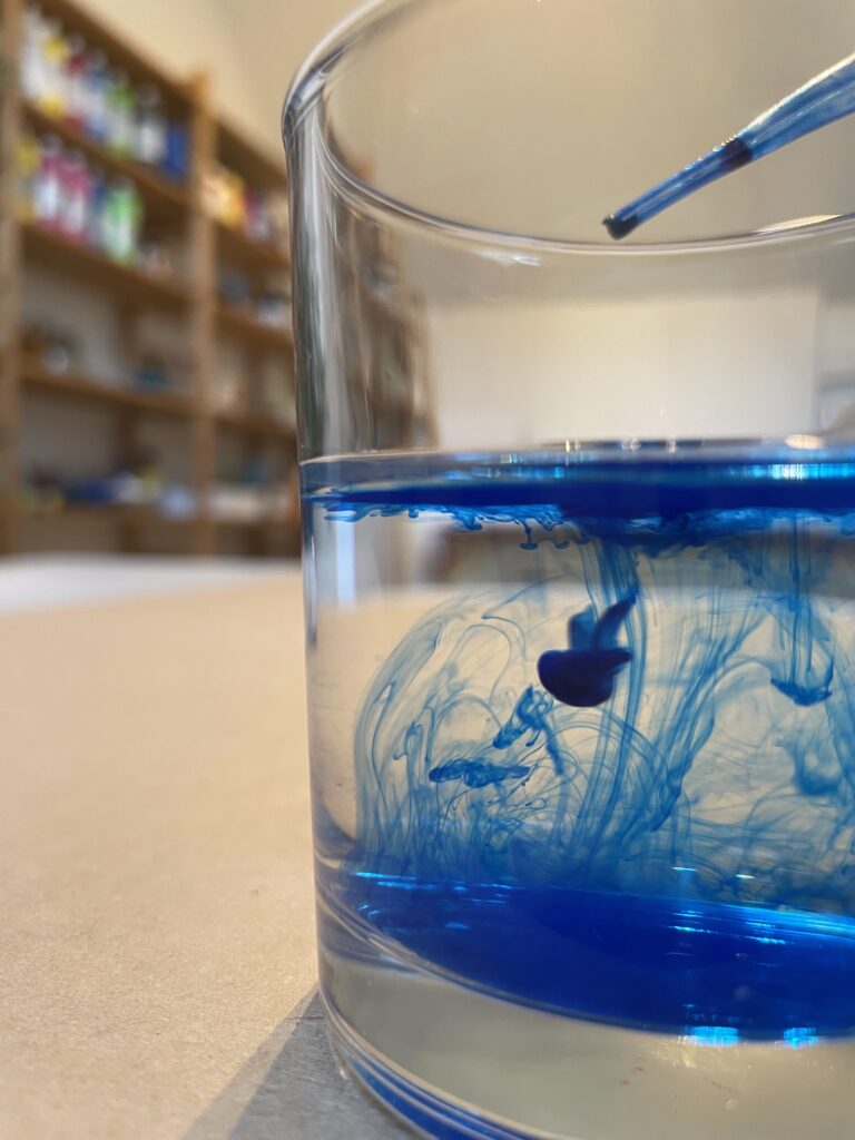 Glas mit Wasser in das ein Tropfen mit blauer Tusche hineingetropft worden ist in einem Kunsttherapie Atelier