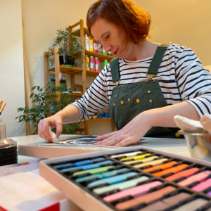 Frau gestaltet ein Bild im Kunsttherapie Atelier mit Pastellkreiden. Im Vordergrund ist eine Farbpalette mit unterschiedlichen Pastellkreiden. Hinter der Frau sind grüne Pflanzen und ein Holzregal