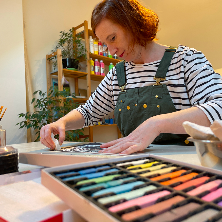 Frau gestaltet ein Bild im Kunsttherapie Atelier mit Pastellkreiden. Im Vordergrund ist eine Farbpalette mit unterschiedlichen Pastellkreiden. Hinter der Frau sind grüne Pflanzen und ein Holzregal